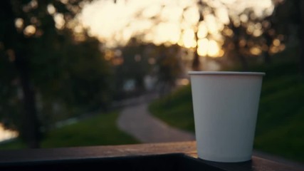 A white paper cup with coffee or tea stands on the railing against the backdrop of the city park at sunset. A hot drink raises steam. - Powered by Adobe