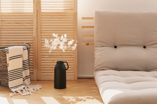 Basket with blanket and white flowers next to grey futon in natural living room interior. Real photo