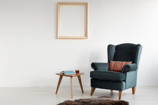 Empty wooden frame on white wall of stylish living room with comfortable armchair with patterned pillow, dark fury carpet and small table with book and tea