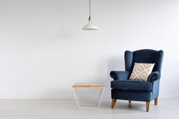 White simple lamp above wooden coffee table in stylish minimal interior with dark blue armchair with patterned pillow, real photo with copy space