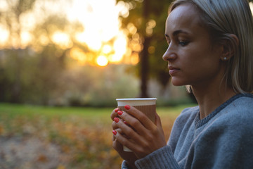 Beautiful young woman drinking hot drink from disposable paper cup outdoors in pretty autumn foliage.