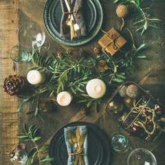 Christmas or New Years celebration party table setting. Flat-lay of plates, golden cutlery, glasses, festive branch decoration, candles and gliterring toys over wooden table background, top view