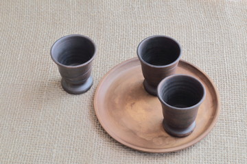 Clay dark brown wine glasses and a round earthen plate on a rough homespun background.