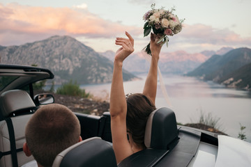 Back view of bride and groom in cabriolet car with hands raised up against the sea