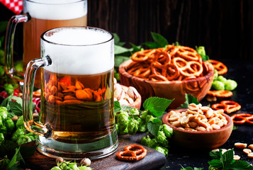 Light german beer poured into big glass, fresh green hops and bowls with salty snacks and nuts, autumn beer festival concept, dark background, selective focus
