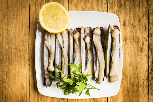 grilled penknives with parsley and half a lemon on a white plate on a wooden table

