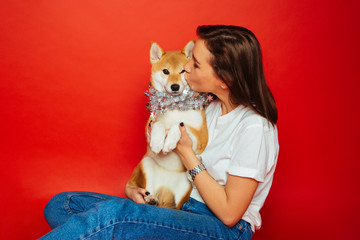 Cute brunette woman in white t shirt and jeans holding and kissing Shiba Inu dog in silver decoration on plane red background. Love to the animals, pets concept