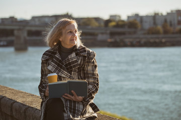 Fototapeta premium Senior woman enjoys reading book and drinking coffee while sitting by the river.