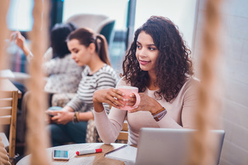 Delicious drink. Positive delighted woman holding a cup with tea while sitting in front of the laptop