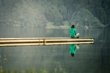 Portrait of young woman in green sweatshirt sitting on the edge of pier and meditating. There is volcanic lake on the background. Peaceful atmosphere. Azores islands, Portugal