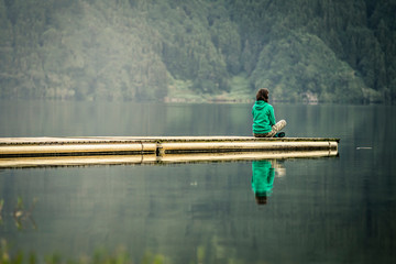 Portrait of young woman in green sweatshirt sitting on the edge of pier and meditating. There is volcanic lake on the background. Peaceful atmosphere. Azores islands, Portugal - Powered by Adobe