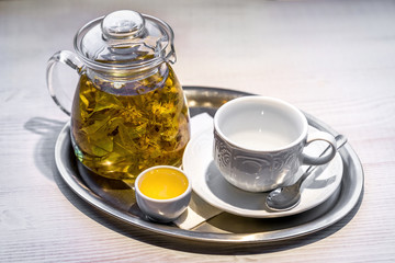 Hot linden-flower tea in transparent glass teapot, small cup of honey, and empty white porcelain cup and silver spoon on saucer arranged on silver tray standing on wooden table in natural light