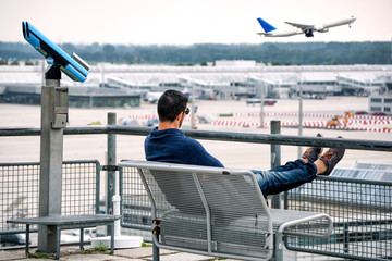Young man sitting on bench watching a plane taking-off at airport.