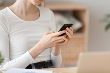 Close up millennial woman sitting at the desk in office room or home holds mobile phone typing messaging email or reading news in internet. Wireless technology, connection online communication concept