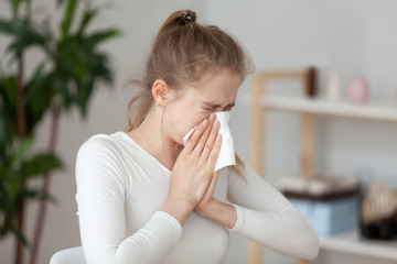 Young millennial sick woman sitting alone at work office sneeze holding tissue handkerchief and...