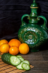 on the table is a vase, cucumbers and tangerines.