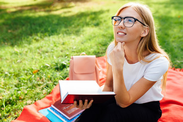 Portrait of a thinking girl student with glasses, sitting on a red blanket in the Park, reading a book and dreaming