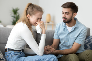 Unhappy married couple on couch at home. Wife crying husband calms her quarrel apologize. Interruption of unwanted pregnancy or miscarriage. Friend support girlfriend after break up with a loved one