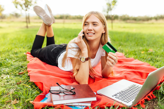 Beautiful business lady, talking on the phone and holding a credit card, sitting on a blanket in the Park