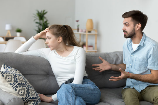Unhappy young millennial married couple sitting on couch in living room at home and quarrelling. Husband arguing emotionally talking frustrated wife looking away. Break up and misunderstanding concept