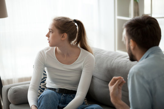 Unhappy couple sitting on couch in living room at home sorting out their relationships quarrelling. Guy arguing emotionally talking frustrated girl looking away. Break up problems in relations concept