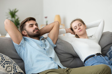 Married millennial couple sitting on sofa in living room at home. Serene tranquil wife husband putting hands behind heads lying and resting with closed eyes. Free time or mindfulness dreaming concept