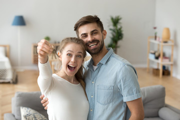 Smiling married couple in love standing hugging in living room at new modern house looking at camera. Excited wife holding keys from their new dwelling. Happy family, moving real estate owners concept