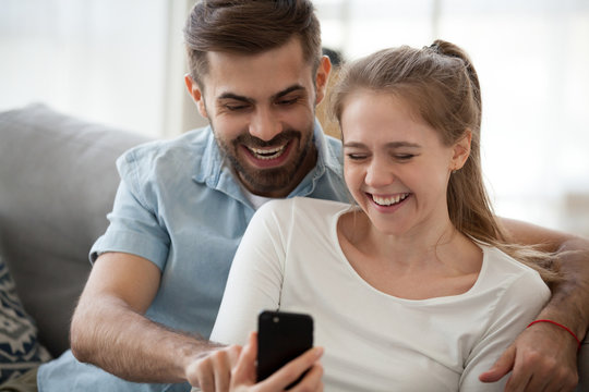 Laughing married couple in love sitting together on couch in living room using mobile phone making selfie photo. Happy husband and joyful wife spend free time on weekend watching funny video online