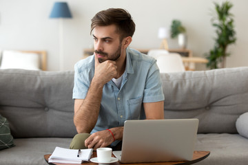 Contemplating millennial serious man looking away sitting on couch. Thoughtful pensive handsome...