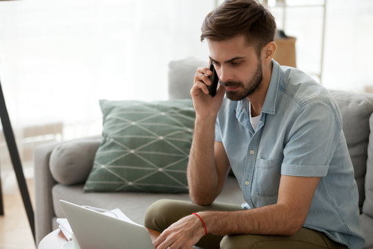 Man sitting on couch reading message email online looking at laptop talking by mobile phone. Male working at home has serious conversation with client. Businessman solve problems and matters by phone