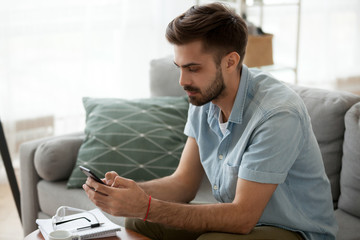 Side view serious man sitting in living room on couch at home chatting typing email using smartphone. Businessman at home solve business matters by phone. New generation addicted with devices concept