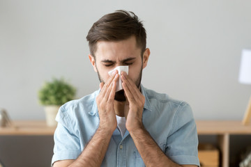 Young sick man sitting at office room sneeze holding tissue handkerchief blowing wiping his running...