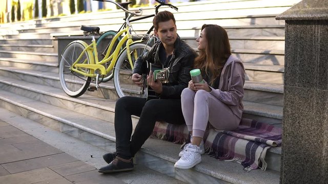Young couple ouple sitting on the stairs and man playing guitar. Man in black casual playing guitar while sitting with his girlfriend outdoors on the city stairs on the plaid. Bikes next to them. Side