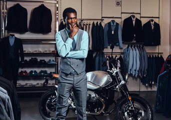 Elegantly dressed African American young man posing near retro sports motorbike at the men's clothing store.