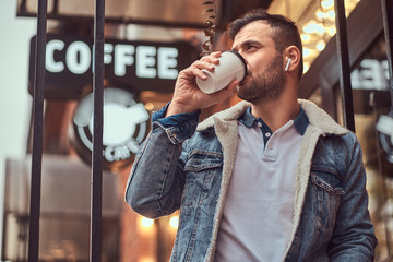 A handsome stylish man wearing a denim jacket with wireless headphones holding takeaway coffee outside the cafe.