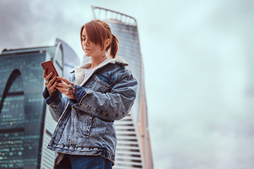 Trendy dressed hipster girl with tattoos on her face and hand using a smartphone in front of skyscrapers in Moskow city at cloudy morning.