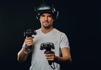 Portrait of a man with bristle dressed in white shirt wearing virtual reality glasses and holds joysticks, looking at camera.