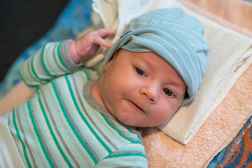 The newborn (2 months baby) with a hat is lying on the bed and laughing