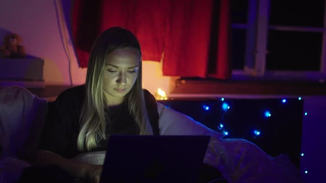 Woman sits on her bed late at night working on a project for work or school. She focuses on the screen while a candle burns behind her in her room.