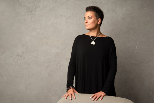 Short-haired woman in a black dress with an amulet around her neck stands leaning against the back of a gray armchair, against the background of a stone wall. Frozen posture of a young beautiful woman