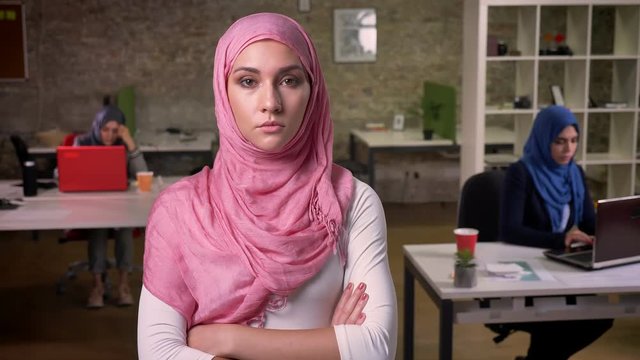 Serious confident nice arab female is standing with hand on hand in pink cute hijab, sitting muslim girls and working on background, modern reality