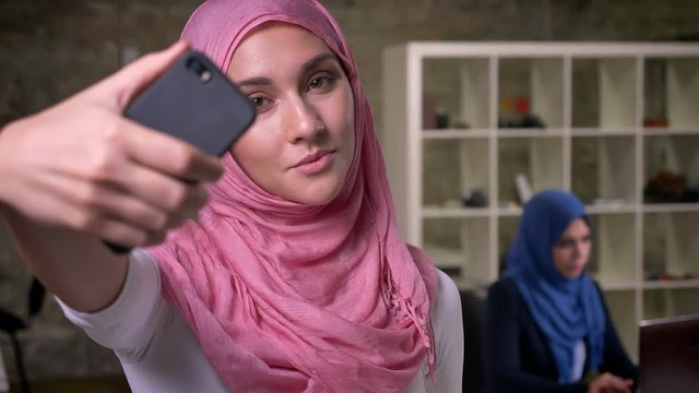 Beautiful focused face of arabic female in pink hijab taking pictures isolated from her working colleagues in brick modern office