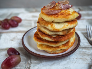 Hill of fresh cheesecakes pancakes on a ceramic plate, ripe grapes and light wooden background