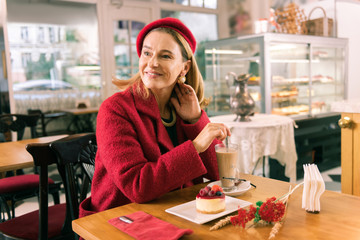 Beaming woman. Beaming mature elegant woman drinking latte and eating dessert in cozy bakery