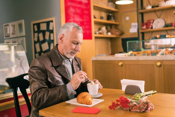 Stirring coffee. Grey-haired man wearing brown leather jacket stirring his coffee spending morning...
