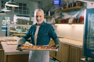 Cinnamon buns. Beaming grey-haired baker feeling satisfied while holding whole tray with nice cinnamon buns