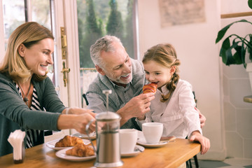 Trying croissant. Cute granddaughter with nice hairstyle trying delicious croissant having breakfast with grandparents