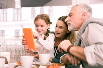 Play on tablet. Dark-eyed granddaughter playing on orange tablet sitting near grandparents