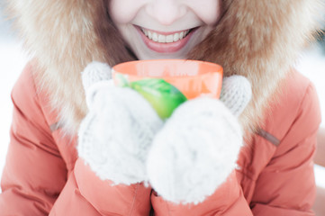 Outdoor portrait of a smiling young girl in a fur hood. Drinking tea from an orange mug in the winter park