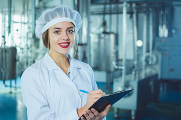 A young beautiful girl in white overalls makes notes in a tablet on the background of equipment of a food processing plant. Quality control in production